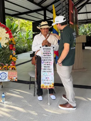 Alvarán conversa con un hombre mayor junto a una silleta de flores, bajo un cobertizo